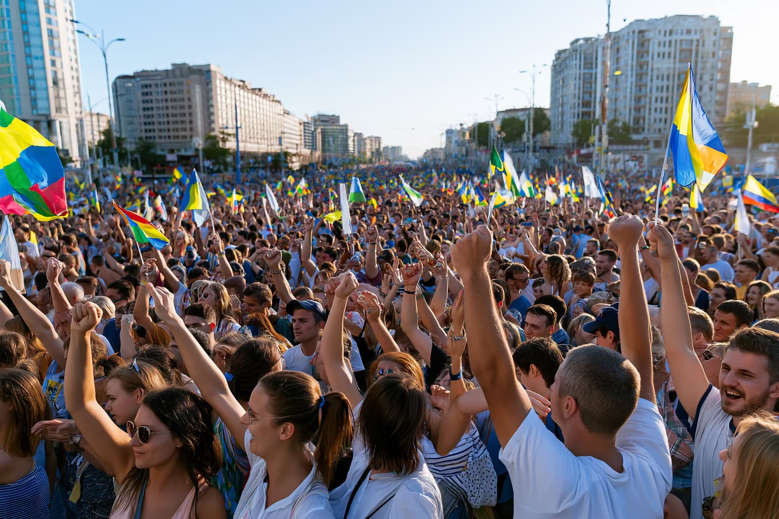 O mulțime imensă umple o stradă a orașului sub un cer luminos de vară, fluturând steaguri colorate.