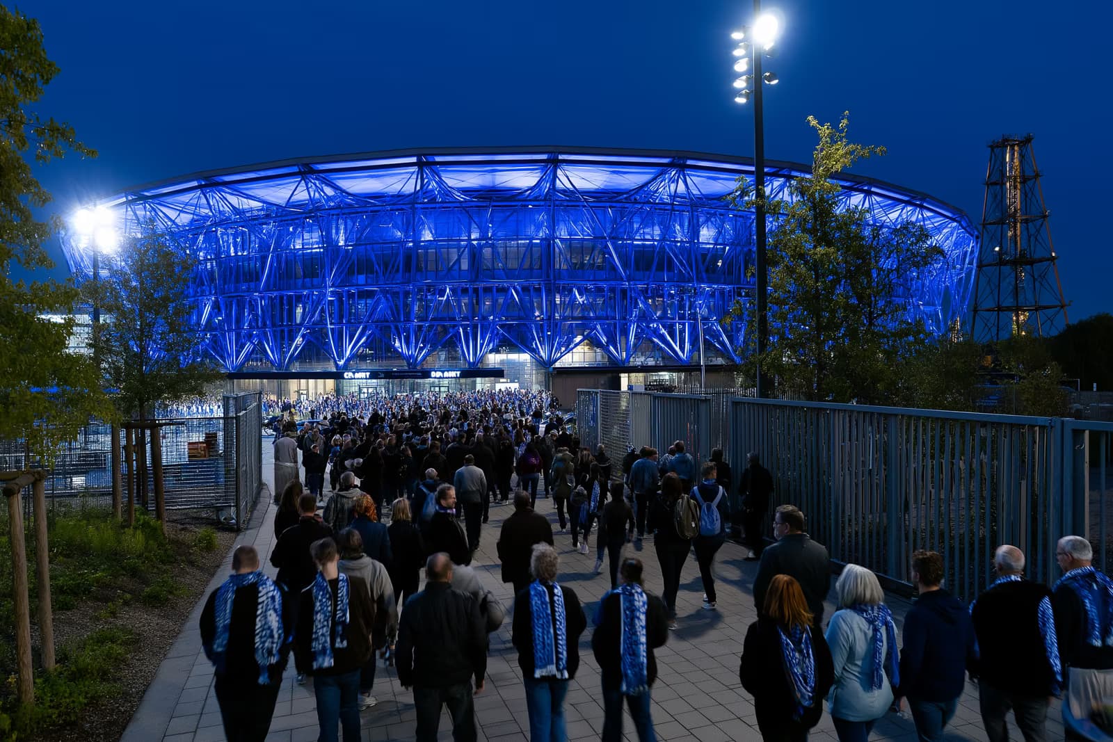 Un stadion de fotbal din zona Ruhr strălucește în albastru regal noaptea, în timp ce fanii cu eșarfe se îndreaptă spre porți după transferul lui Gosens la Schalke.